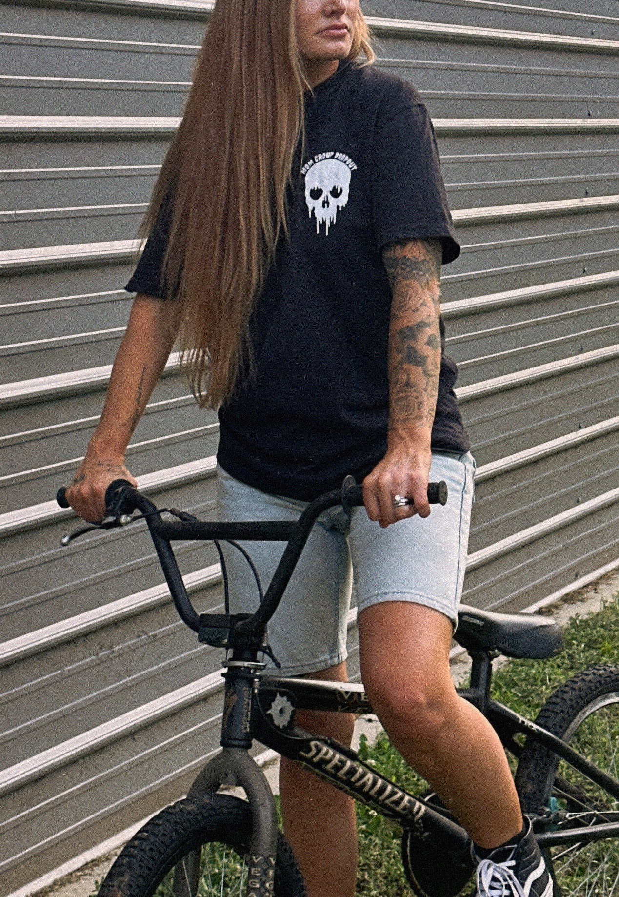 A woman with long light brown hair sits on a BMX bike in front of a corrugated metal wall. She is wearing a black t-shirt with a white skull graphic, light gray denim shorts, and black sneakers.
